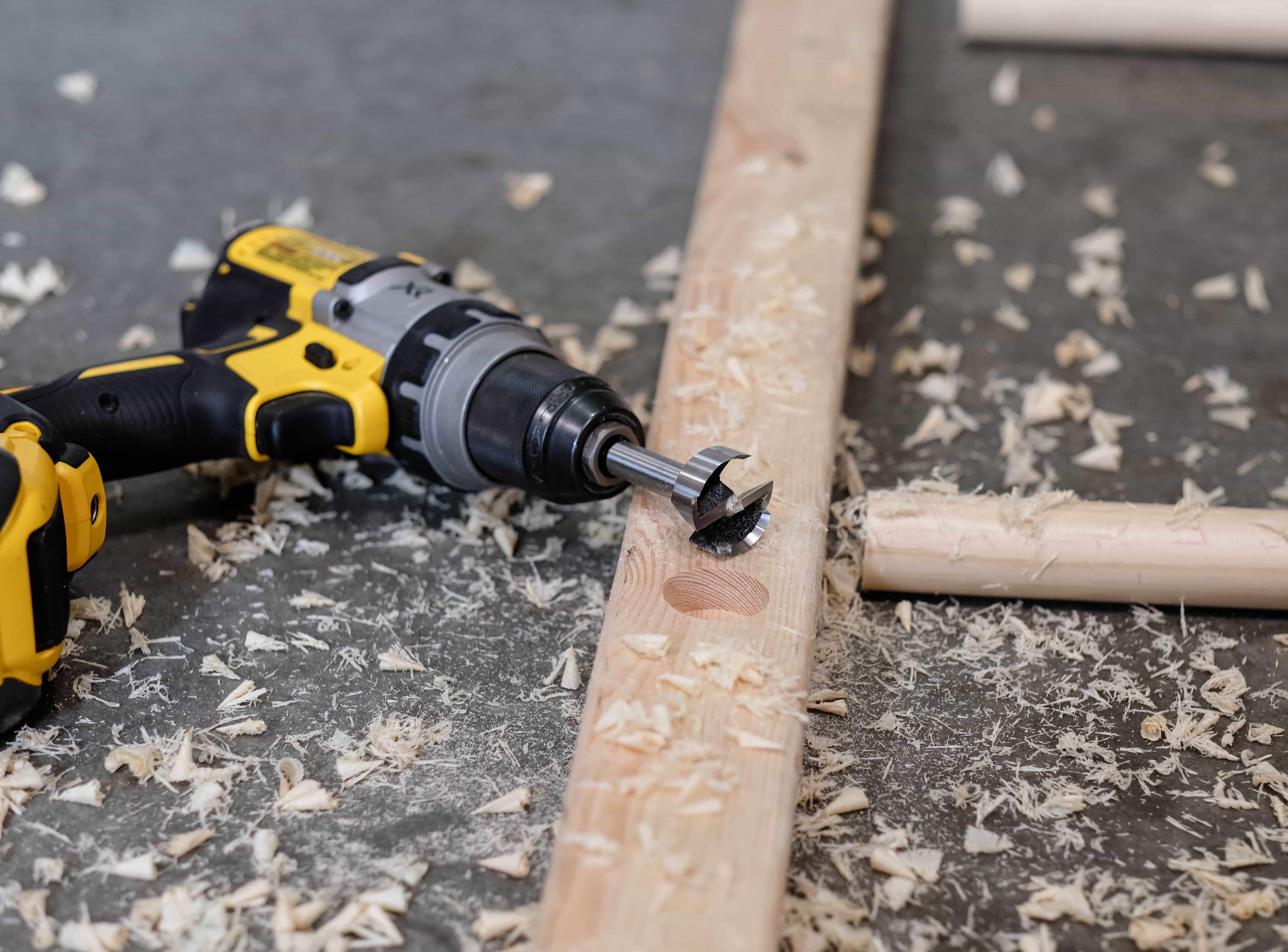 A board with a hole drilled in it. The drill is laying next to the board. The board is for a blanket ladder.