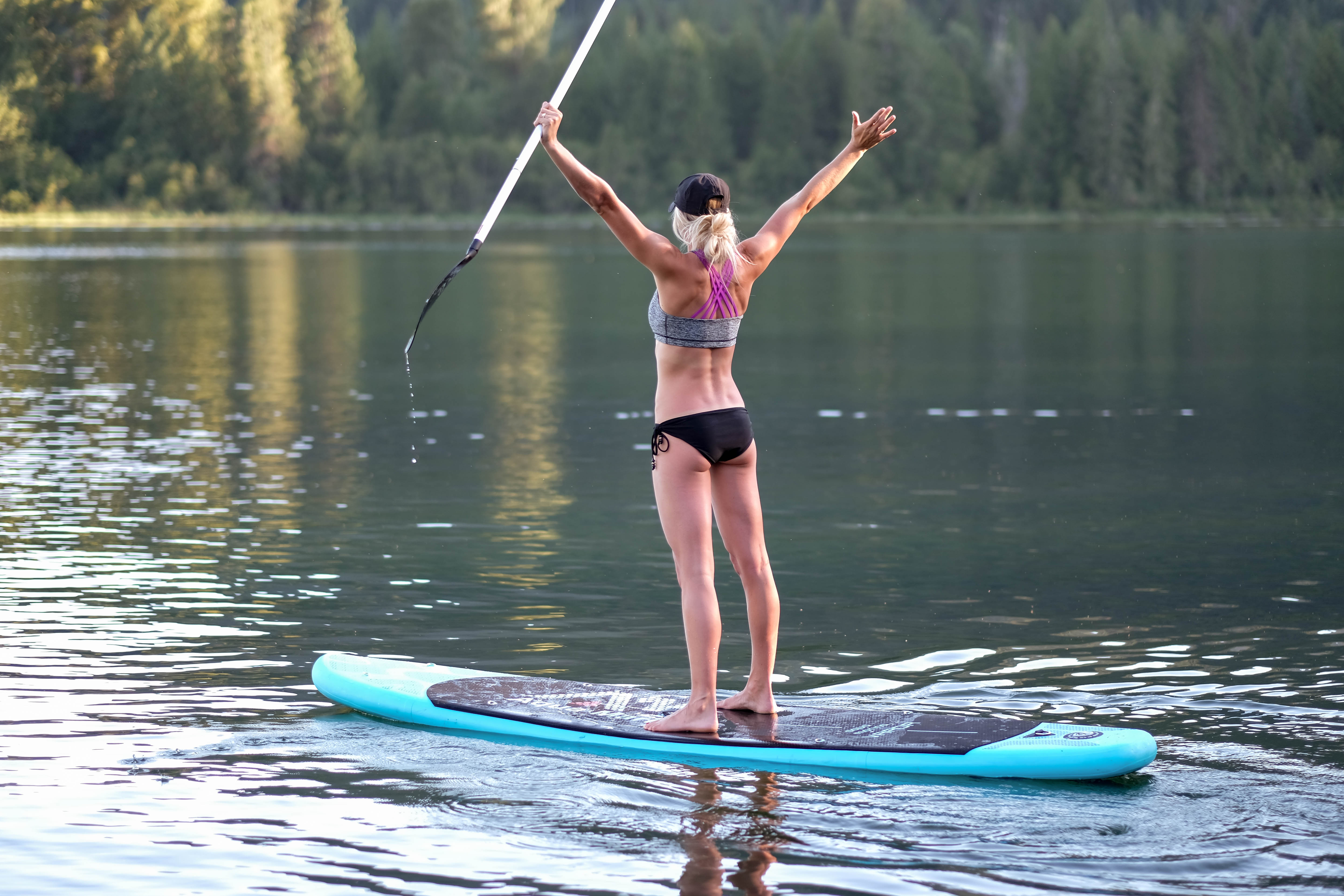 Blond girl standing on inflatable SUP in the middle of a lake with arms raised