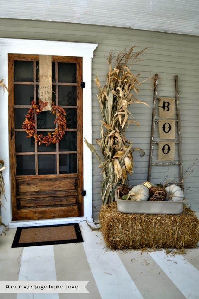 Fall front porch ideas using a hay bale, corn stalks and a rustic blanket ladder with burlap flags that read B-O-O