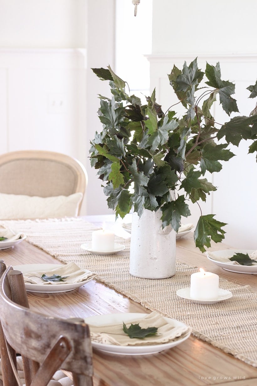 Thanksgiving table centerpieces with a large white pitcher filled with stems with large green leaves on a jute runner